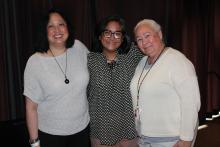 Three women stand shoulder to shoulder smiling in front of a stage curtain