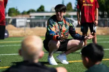 Han Eckelberg wearing a multi-color long-sleeve shirt, crouching in front of a group of children speaking to them