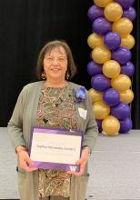 A woman stands in front of a stage holding a white parchment paper that reads Anjélica Hernández-Cordero 2024 distinguished staff award nominee. Behind her is a stack of purple and gold balloons.