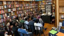 Linh Thủy Nguyễn sits in front of an audience wearing a black face mask speaking about her book Displacing Kinship