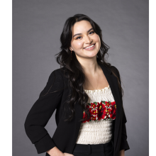 María stands in front of a gray background, smiling, wearing a white shirt with a floral pattern across the middle and a black blazer over the shirt