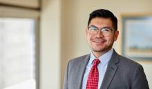 Paulo smiles while standing and wearing glasses, a light blue button-down shirt with a red-tiled necktie, and gray blazer