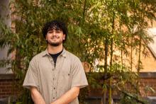 Lorenzo McCleese smiles while standing in front of a bamboo tree wearing a light tan striped button down with a black undershirt