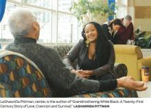 LaShawnDa, centered in the photo, is sitting on a couch wearing a gray shawl over a black shirt and is talking with an African American grandmother
