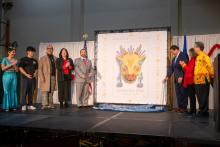 Seattle Mayor Bruce Harrell pulls a string to reveal the new Year of the Dragon Forever stamp at the International District Community Center. There are five individuals standing stage right and three standing stage left. An enlarged image of the Year of the Dragon stamp center stage.