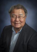 Dr. Stephen H. Sumida smiles in front of a gray background wearing glasses, a dark gray suit jacket, and a blue buttoned-down shirt