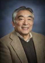 Tetsuden Kashima smiles in front of a blue background and wearing a light gray blazer with a dark gray sweater underneath