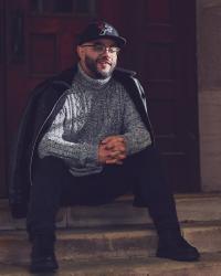 Alekzandr Wray sits on a short staircase in front of a door with his hands clasped  wearing a black hat with a rose cutting through a cross, glasses, a black jacket draped over his shoulders, and a gray, white, and black sweater