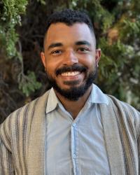 Matthew Randolph smiles while standing in front of some greenery wearing a light blue button-down shirt with a tan with black stripes sweater over