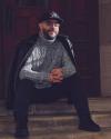 Alekzandr Wray sits on a short staircase in front of a door with his hands clasped  wearing a black hat with a rose cutting through a cross, glasses, a black jacket draped over his shoulders, and a gray, white, and black sweater