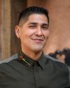 Lupe smiles wearing a black button down shirt with gray stripe across the shoulder and a rosary lapel pin on the right collar