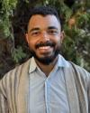 Matthew Randolph smiles while standing in front of some greenery wearing a light blue button-down shirt with a tan with black stripes sweater over