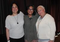 Three women stand shoulder to shoulder smiling in front of a stage curtain