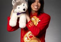Dylan Lai smiles while holding up a stuffed Husky on his right shoulder, wearing traditional red garments with a gold dragon sprawling vertically