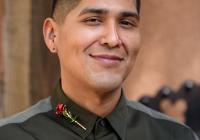 Lupe smiles wearing a black button down shirt with gray stripe across the shoulder and a rosary lapel pin on the right collar