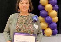 A woman stands in front of a stage holding a white parchment paper that reads Anjélica Hernández-Cordero 2024 distinguished staff award nominee. Behind her is a stack of purple and gold balloons.