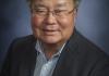 Dr. Stephen H. Sumida smiles in front of a gray background wearing glasses, a dark gray suit jacket, and a blue buttoned-down shirt