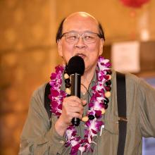 An Asian man wearing an olive button-down shirt, black suspenders, a lei adorned with pink and white flowers, a necklace with beads, speaks to the audience using a microphone held in his right hand