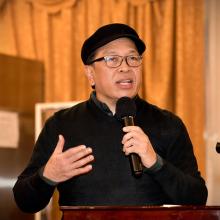 An Asian man wearing a black hat, black sweater over a gray button-down shirt, and glasses, speaks to the audience using a microphone in his left hand